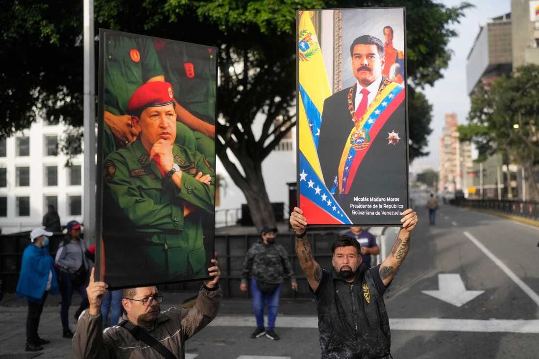 Two men in downtown Caracas hold up large, framed portraits of Venezuelan leaders. The man on the left holds a portrait of Hugo Chávez in military fatigues and a red beret. The man on the right lifts a portrait of Nicolás Maduro wearing the presidential sash high above his head.