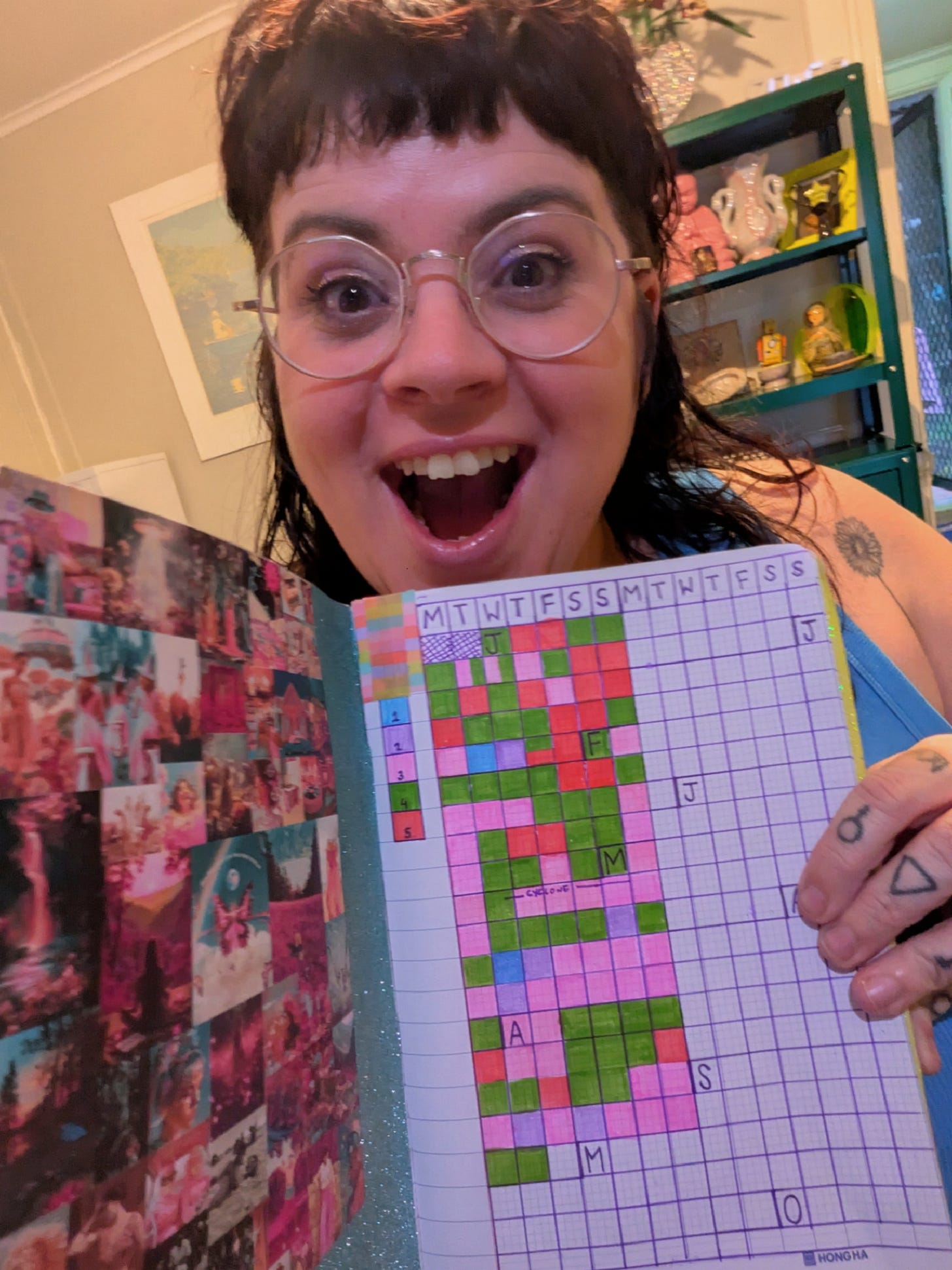 A selfie of Claire, a plus size white woman, with dark hair and a fringe, tattoos and gold glasses, holding a notebook with coloured in squares. 