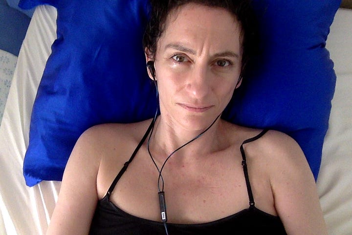 On the left, a girl with brown hair and watery eyes looks pained or concerned. She wears a black top, black wired headphones, and lays on a bright blue pillowcase. On the right, the same girl is smiling, lying sideways on her bed, wearing a bun and a green tank top in front of a brown slatted headboard.