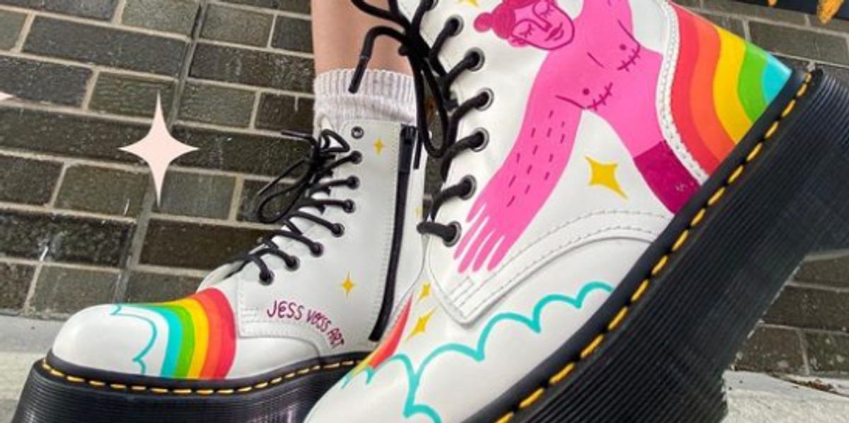 A photo of someone modeling a pair of white Doc Marten boots that feature rainbows on the toe and a pink illustration of a person with top surgery scars on the side of the shoe.
