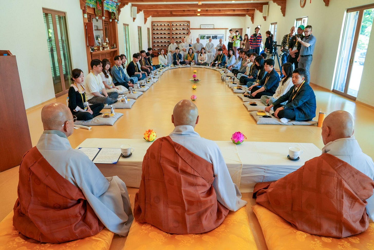 A large group of participants sit on floor cushions arranged in a wide semi-circle inside a temple hall with three monks at the front.