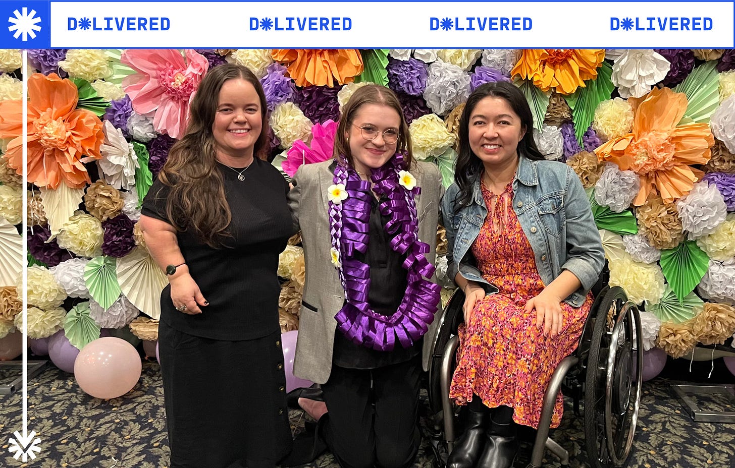 Ella, Annabel and Olivia smile warmly in front of a colourful wall made of paper flowers