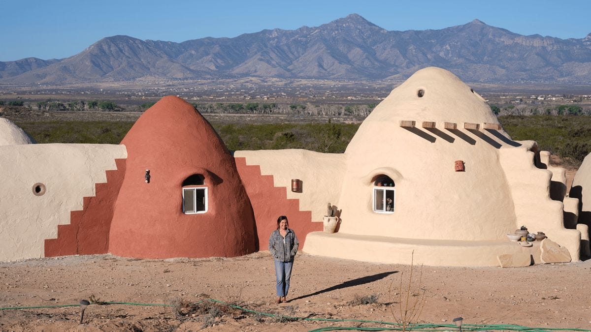 A person stands in front of two dome-shaped adobe houses, embracing homesteading life in Cochise County, with mountains rising in the background under a clear sky.