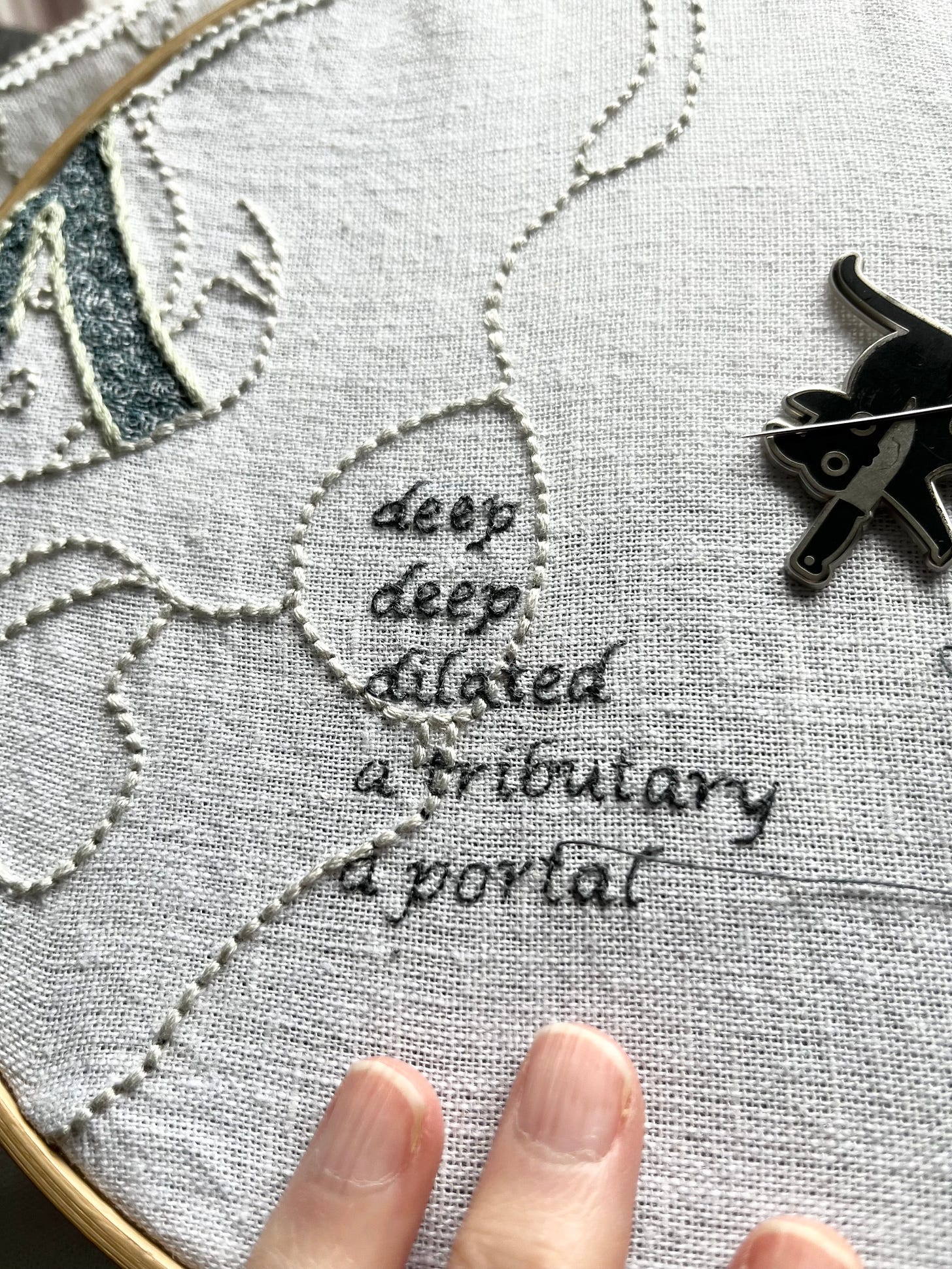 The left side of a pelvis, lumbar vertebrae, and part of the iliac vein embroidered on white linen with some of it in a bamboo hoop. Most of the iliac vein is outlined in grey-green stem stitch. A section on the left branch is outlined in darker grey-green split stitch with honeycomb filling stitch stretched across it. Intricate turbulence is stitched in single strand dark aquamarine backstitch within the iliac vein. The bones are stitched in thick backstitch in a white just slightly bluer than the white linen. A block of text is stitched in dark grey with some letters thicker and smoother than others. It reads: Deep. Deep. Dilated. A tributary. A portal. A black knifecat needle minder holds the needle.