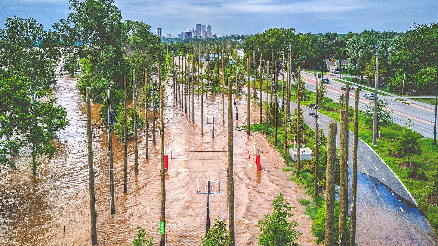 Mud-orange floodwaters lap across the sports courts at Gathering Place beneath numerous wood-clad light poles, bright green vegetation and a red volleyball net. The water spills across a portion of the paved trail as cars pass on Riverside Drive. Mud-orange floodwaters lap across the sports courts at Gathering Place beneath numerous wood-clad light poles, bright green vegetation and a red volleyball net. The water spills across a portion of the paved trail as cars pass on Riverside Drive.