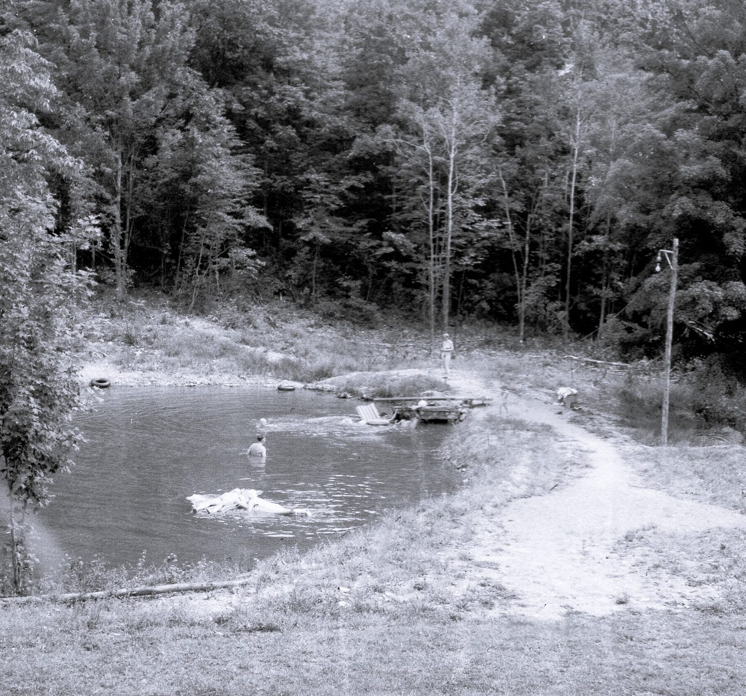A small pond at a wooded nudist camp, with a nude swimmer in the water and a few people standing along the path by the shoreline.