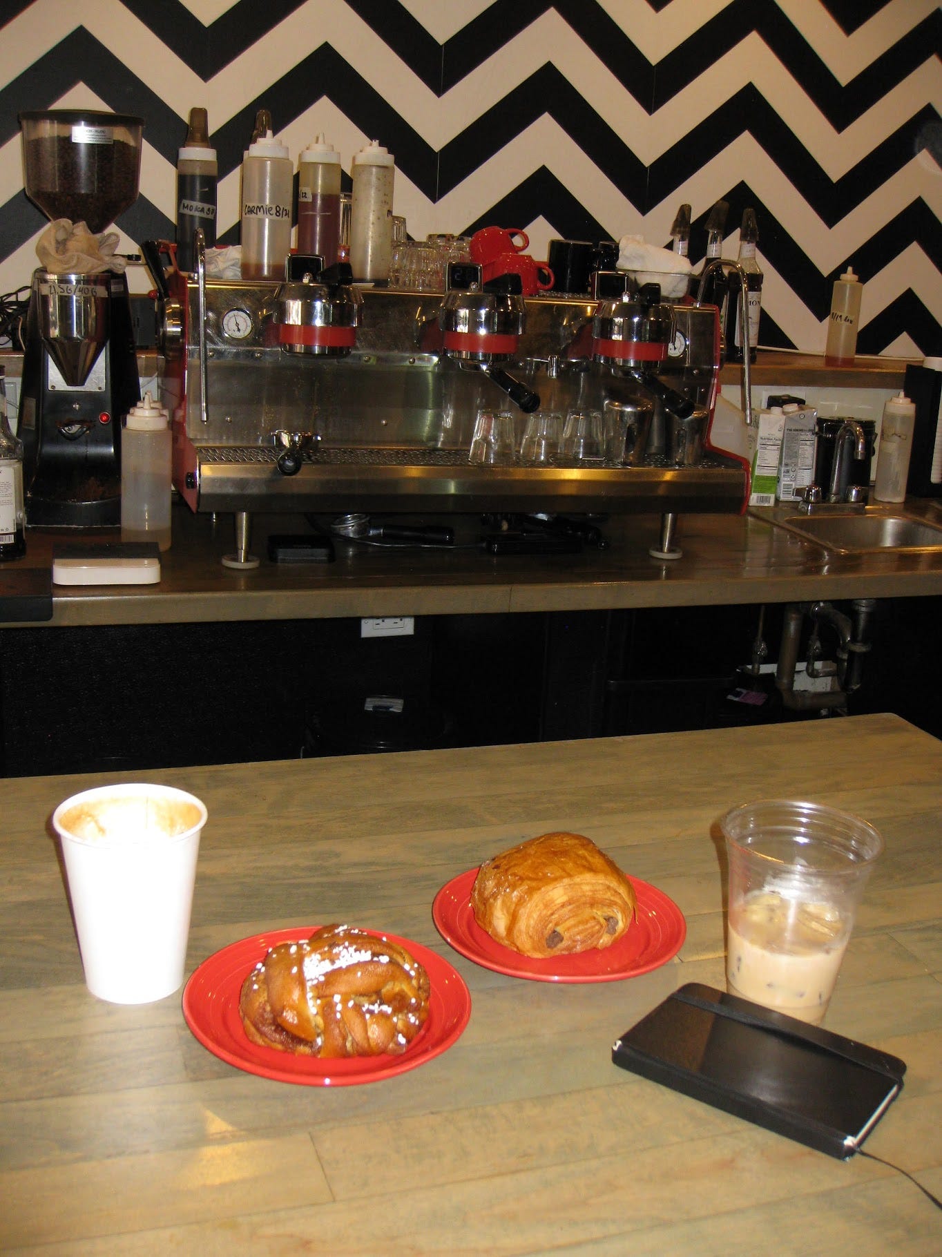 The background of this photo is an expression machine with various syrups and sweeteners on top of it. The wallpaper is black and white chevorn. In the foreground, there are two red ceramic plates: one with a chocolate crossaint and the other with a braided bread. On either side of the plates are drinks, one iced and one hot.