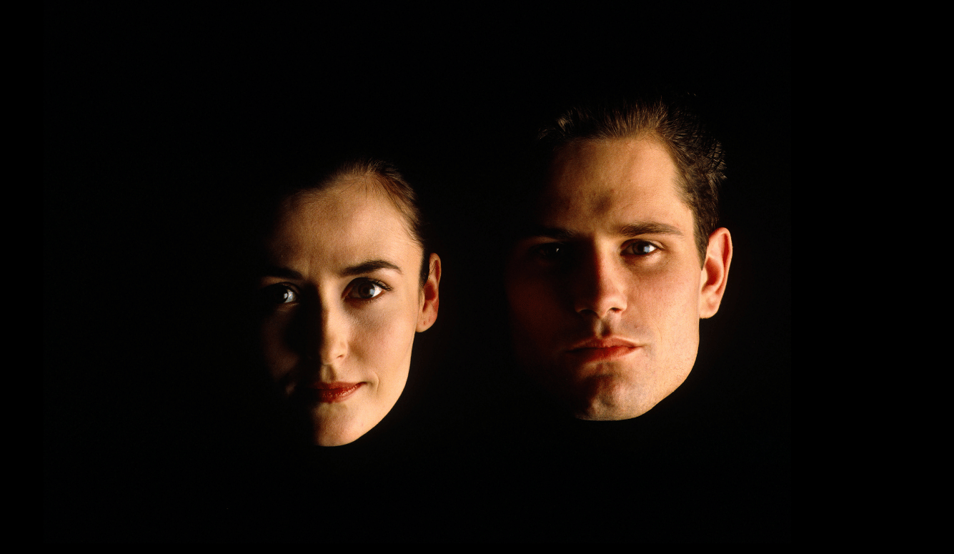 Two faces—one woman on the left and one man on the right—are lit dramatically against a black background, with the rest of the image in darkness.
