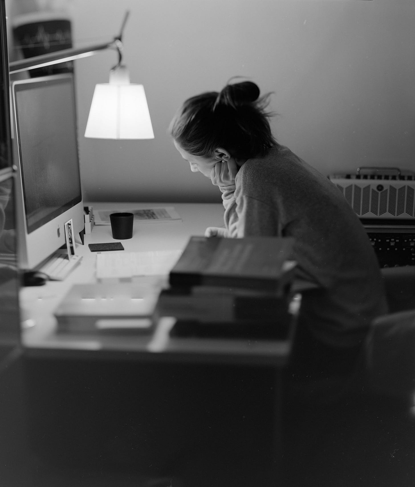 A black-and-white photo of a woman seated at a desk, leaning on her elbow, in front of a large computer screen. A stack of books rest on the desk in the foreground, and a small overhead light hangs over the desk surface.