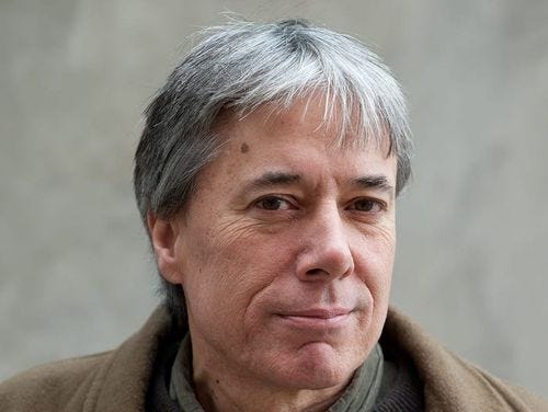 Headshot of Chuck. 60 something white man with grey and black medium length hair, a half smile, looking directly into the camera. Brown eyes and a brown coat on.