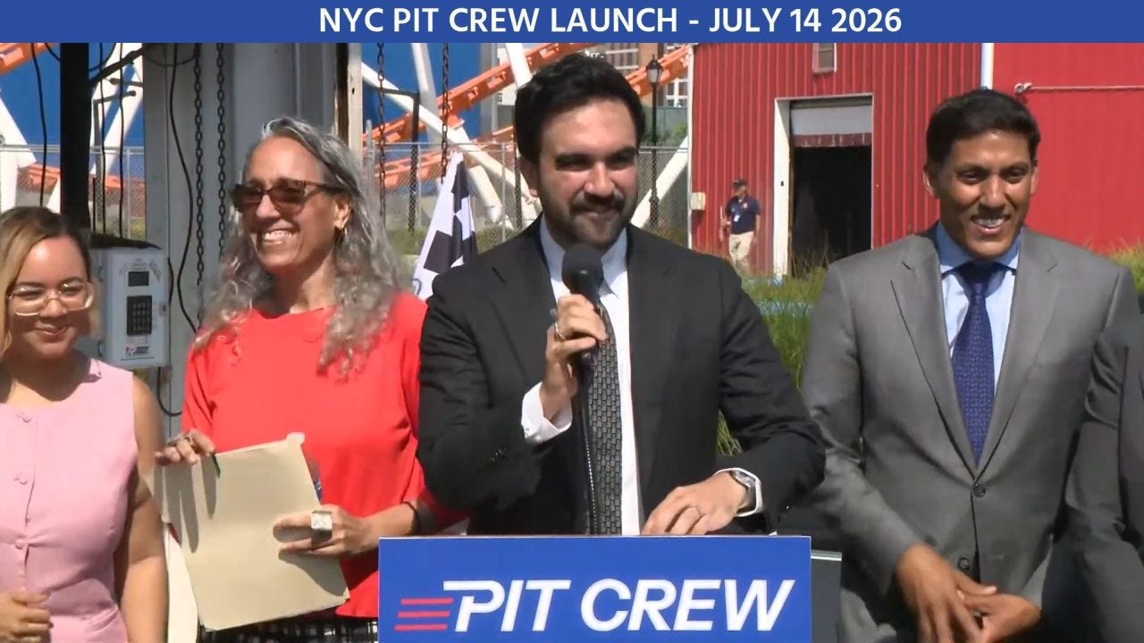 A wide outdoor event scene showing several people standing together behind a blue podium at what appears to be an amusement park or industrial venue. A roller coaster track and structural supports rise in the background alongside a red corrugated building, while bright midday sunlight casts strong shadows. A blue banner spans the top of the image, and the podium carries matching blue branding, creating a coordinated event look. The speaker at center holds a microphone while those beside him smile, giving the image the feel of a ceremonial announcement or press event.