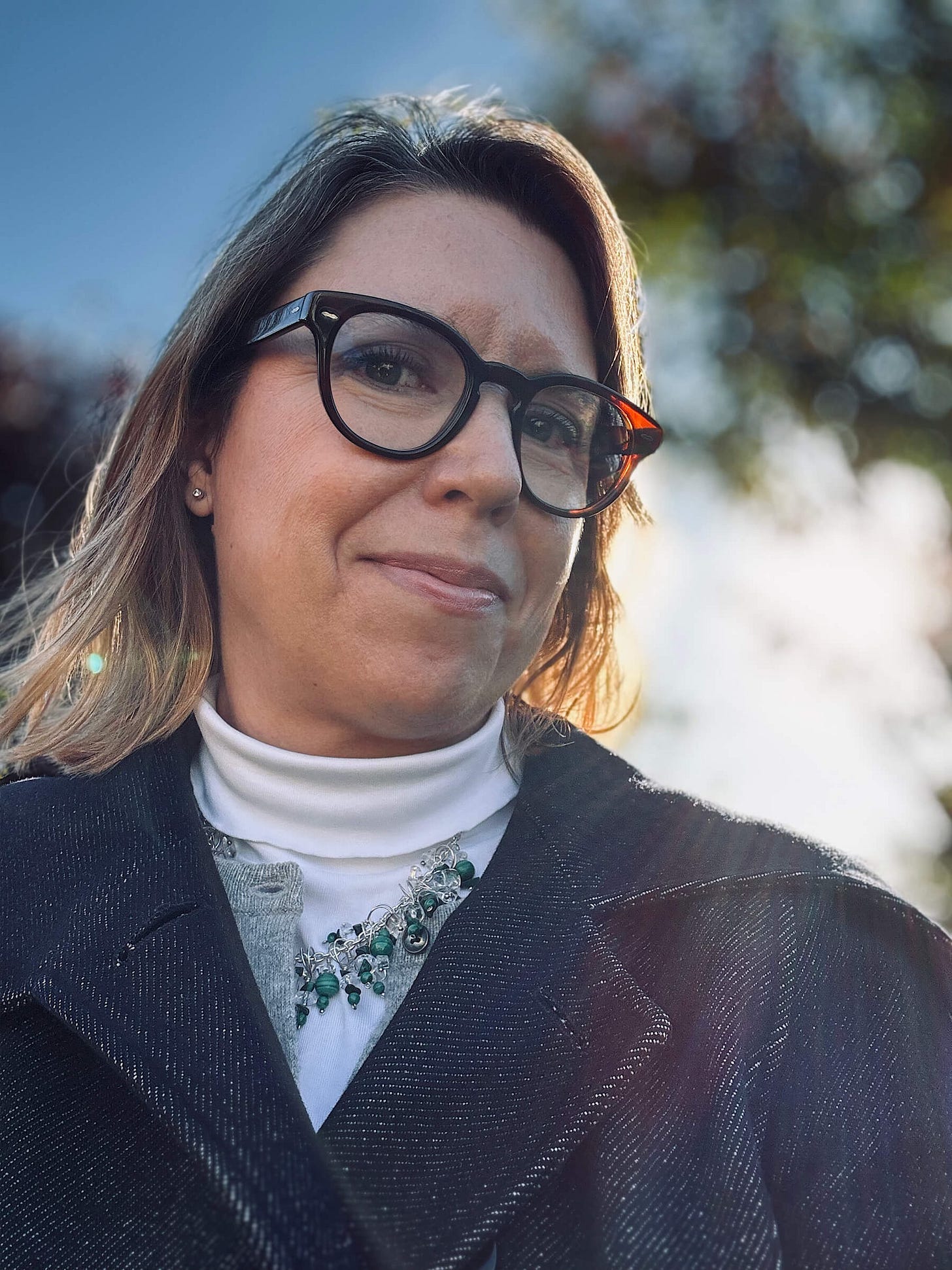 woman standing with the sun behind her wearing a blue coat, grey cardigan, beaded necklace and brown rimmed glasses
