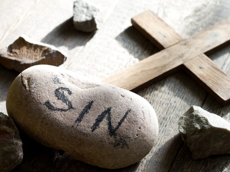 A rock with the word "sin" on it rests beside a small wooden cross and smaller rocks.