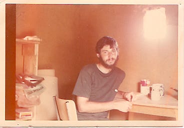 Photo of a 22 year old man wearing a t-shirt sitting at a simple tale inside a small adobe building. Light comes in through an un-glazed window.