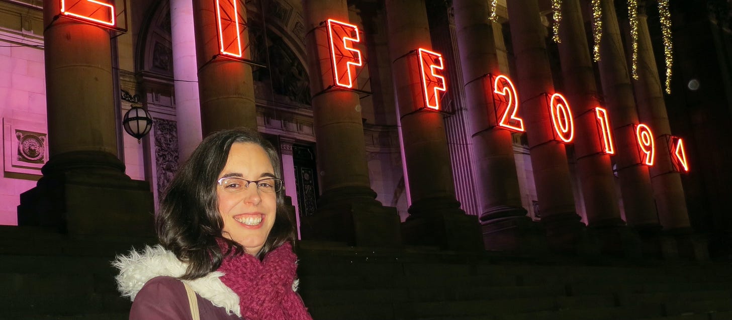 Headshot of Vicki in a winter coat and scarf in front of a pink neon sign saying LIFF 2019