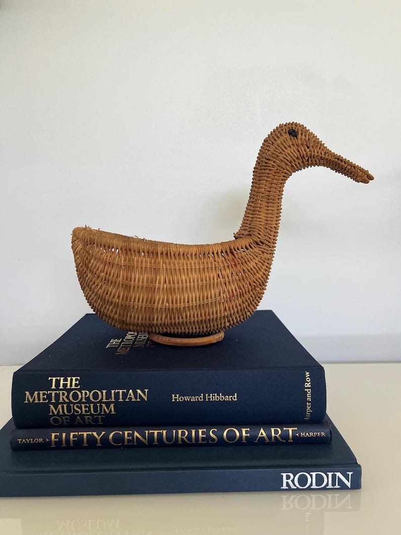May include: A woven wicker duck shaped basket with a brown finish. The basket is sitting on top of three navy blue books. The books are titled 'The Metropolitan Museum of Art Fifty Centuries of Art' and 'Rodin'.