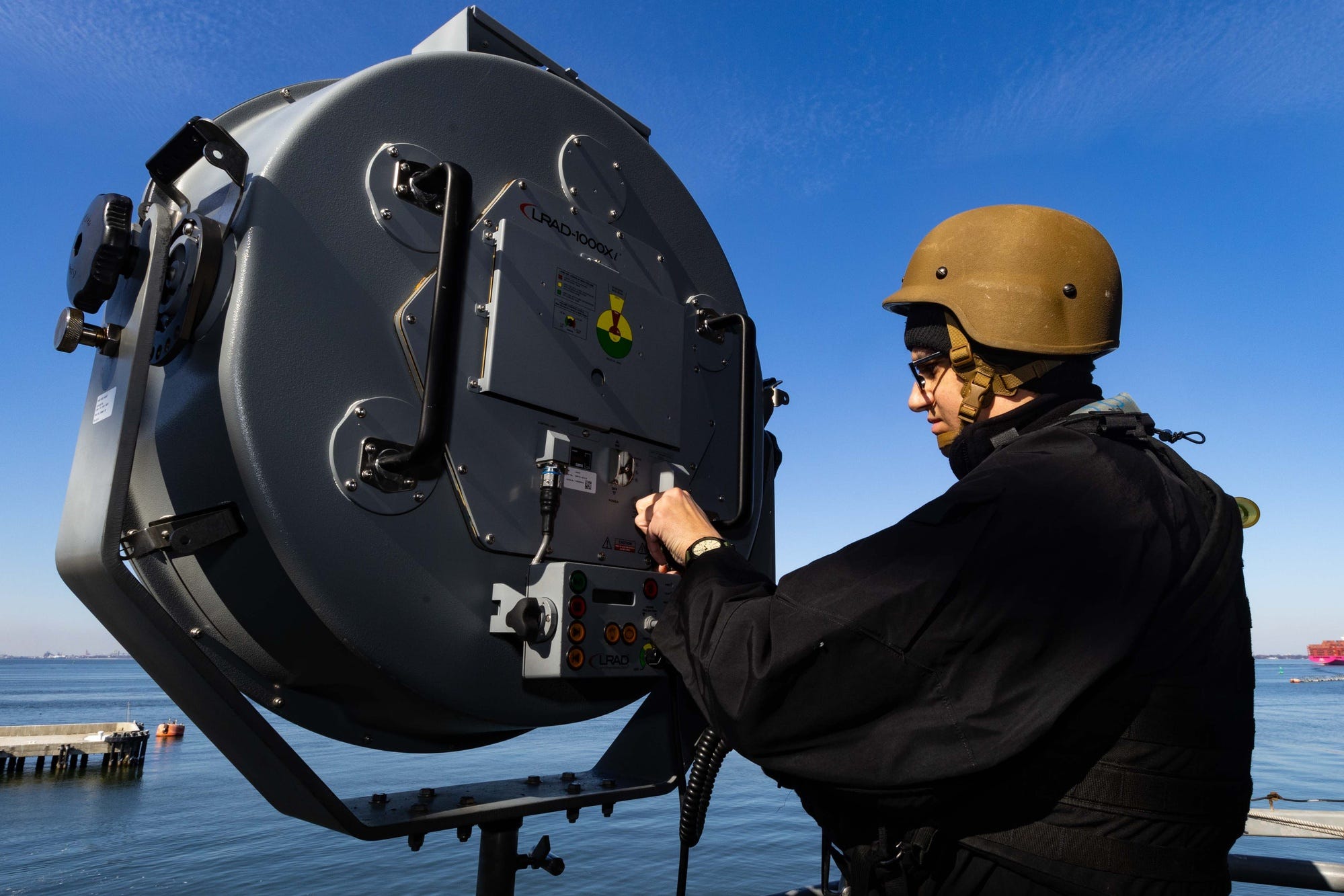 Helmeted sailor operates the rear controls of a large circular gray LRAD aboard USS Gerald R. Ford beneath a clear blue sky.