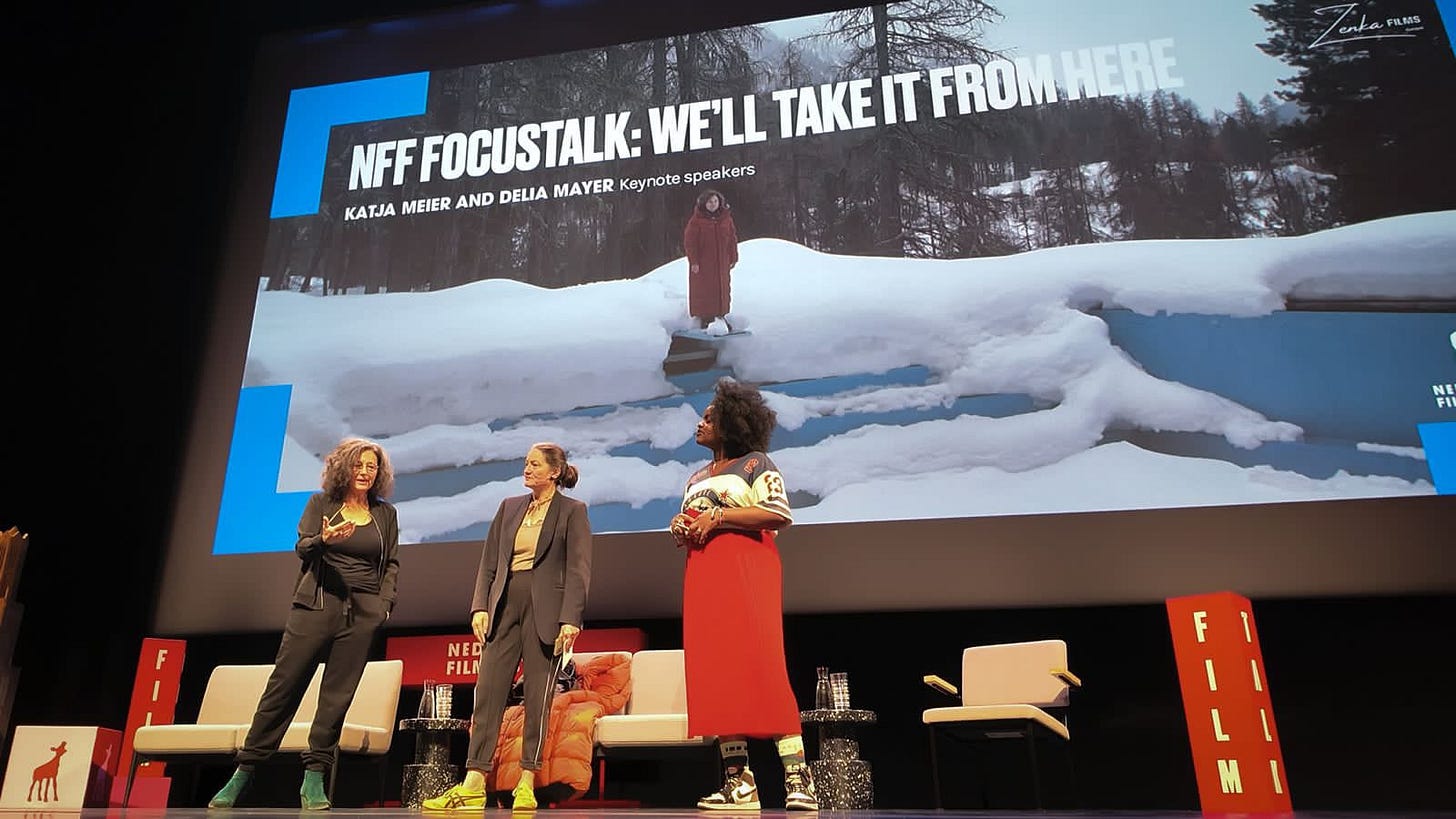 three women on stage with a big screen in the back that shows the film poster image of the TV series they talk about (a woman standing a top of a snow-covered pool)