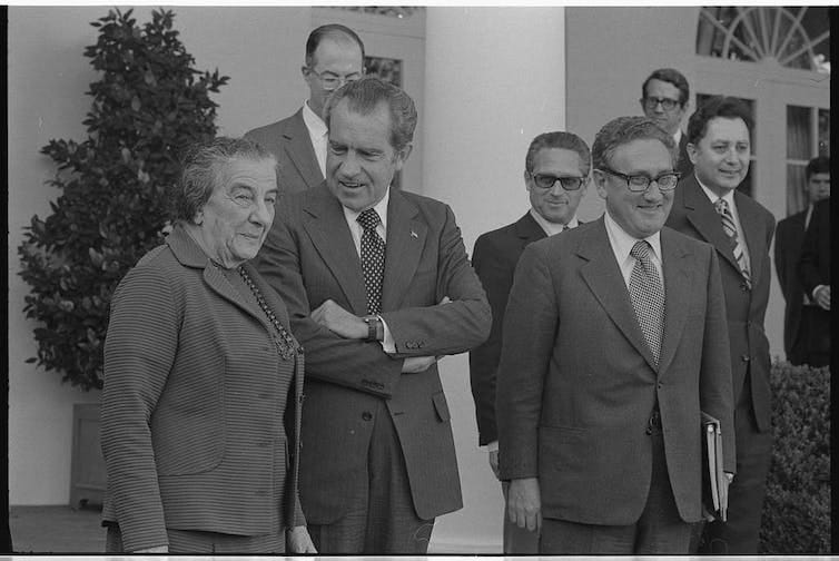 Henry Kissinger, Richard Nixon and Golda Meir stood smiling with aides. Henry Kissinger, Richard Nixon and Golda Meir stood smiling with aides.