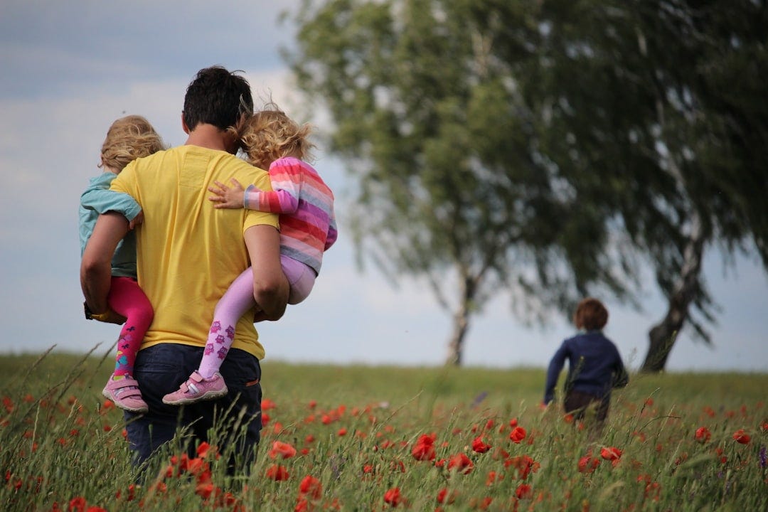 man carrying to girls on field of red petaled flower man carrying to girls on field of red petaled flower