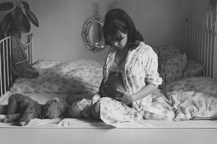 This may contain: a pregnant woman sitting on the bed with her teddy bear and looking down at her belly