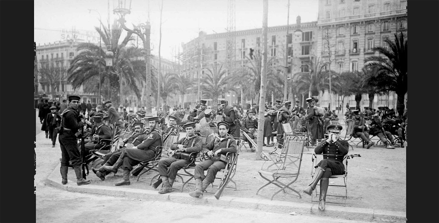 Fotografia storica in bianco e nero scattata nel 1919 a Barcellona nella quale si vedono militari seduti a riposo su sedie in ferro battuto in una piazza decorata con delle palme. Sullo sfondo si vedono edifici. Fotografia storica in bianco e nero scattata nel 1919 a Barcellona nella quale si vedono militari seduti a riposo su sedie in ferro battuto in una piazza decorata con delle palme. Sullo sfondo si vedono edifici.