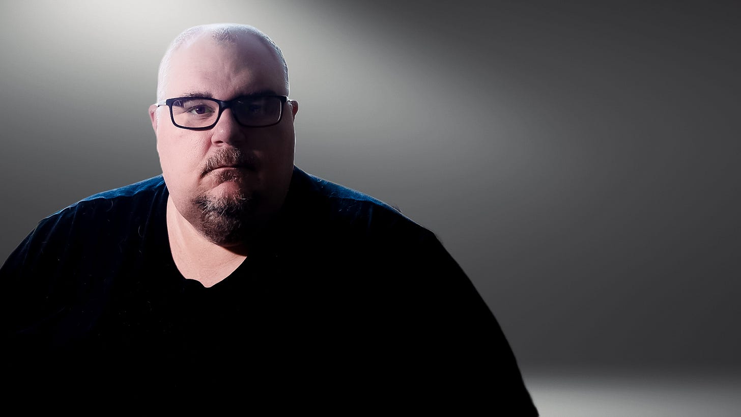 A serious-looking man with glasses and a goatee, wearing a dark shirt, sitting in moody, focused lighting against a neutral background.