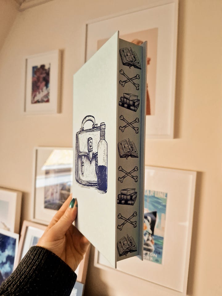 L: the naked hardback of Bookshops and Bonedust from The Broken Binding. It's pale blue with a darker blue embossed image of a satchel and a vial. R: The same book held up to show the sprayed edges, alternating pictures of a pile of books, two crossed bones, and an open book.