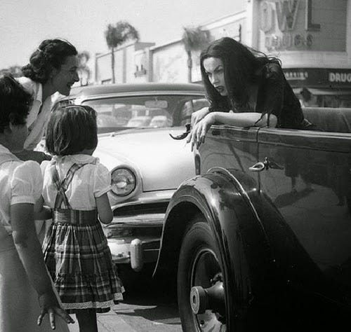 A gothic woman with long black hair and long nails leans out of a hearse to talk to a couple of eager children and their parent