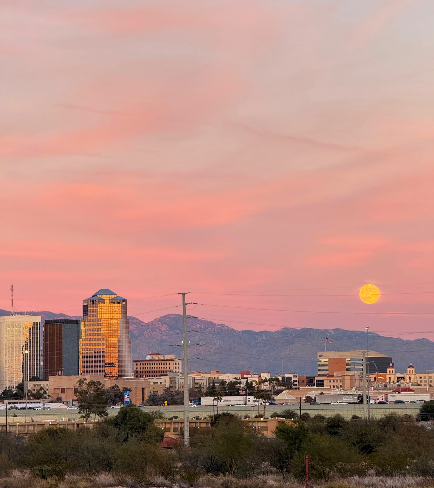 An orange moon rises into a pink sky, over blue mountains and the skyline of Tucson, Arizona.