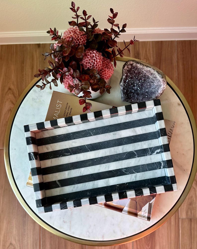 May include: A decorative tray with black and white stripes, resting on a marble-topped table. The tray is rectangular with a striped border. A vase of pink flowers and a geode are also on the table, creating a stylish home decor scene.