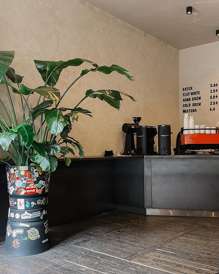 Image 1: Trendy café interior with lime-washed walls, black furniture, and a striking red coffee machine, alongside a large banana leaf plant. / Image 2: Table shot with a glass of water, cup of coffee, a canelé, and my current read, framed by the banana tree in the background.