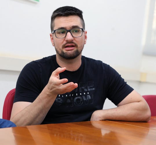 Pessoa usando óculos de armação escura e camiseta preta com estampa, sentada à frente de uma mesa de madeira clara com as mãos gestualizando enquanto fala, em ambiente interno com parede branca ao fundo e cadeira vermelha parcialmente visível. Pessoa usando óculos de armação escura e camiseta preta com estampa, sentada à frente de uma mesa de madeira clara com as mãos gestualizando enquanto fala, em ambiente interno com parede branca ao fundo e cadeira vermelha parcialmente visível.