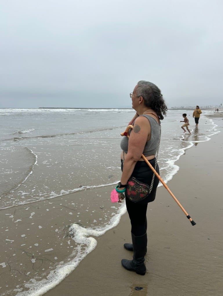 image of the author looking at the ocean in San Diego, grey overcast, in grey crop sweater black miniskirt and tights, pink KN94 around wrist and cane under armpit