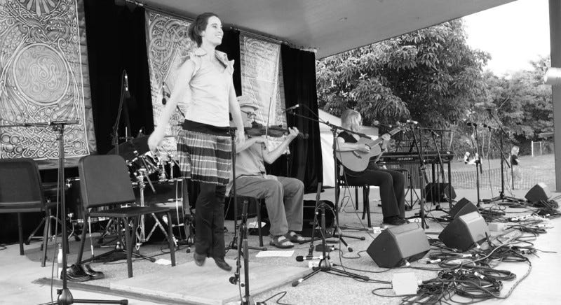 Female step dancer on stage with seated fiddler and guitarist.