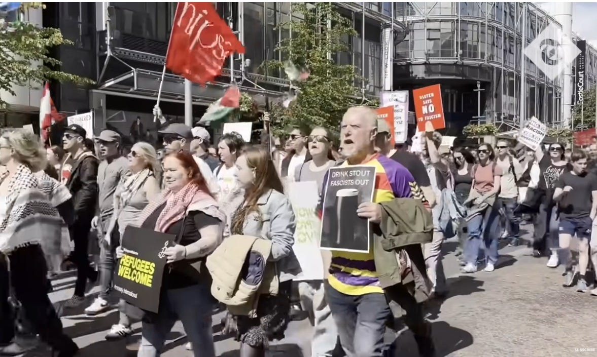 video screenshot of anti-racist march in Belfast. Signs read 'refugees welcome,' 'Drink stout / fascists out' (with photo of pint of Guinness), 'Ulster says NO to fasscism,' and 'No to racism, yes to diversity.' Most marchers are white, as in other UK counter-protests to racist mobs. video screenshot of anti-racist march in Belfast. Signs read 'refugees welcome,' 'Drink stout / fascists out' (with photo of pint of Guinness), 'Ulster says NO to fasscism,' and 'No to racism, yes to diversity.' Most marchers are white, as in other UK counter-protests to racist mobs.