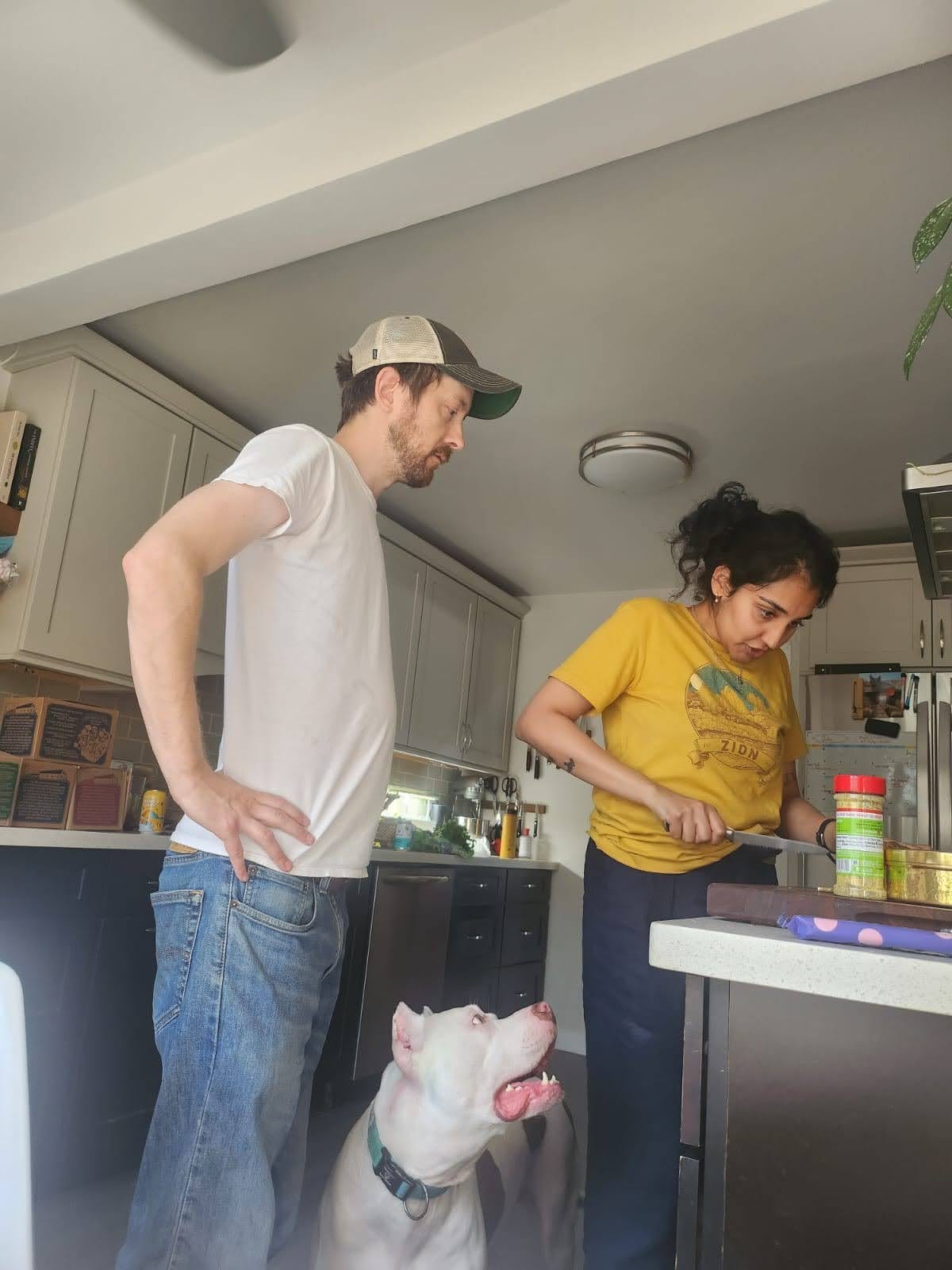 Man on left, standing with his hands on his waist. The woman on the right is cutting something on the cutting board. The white dog standing in the middle is looking at the woman, expecting some food.