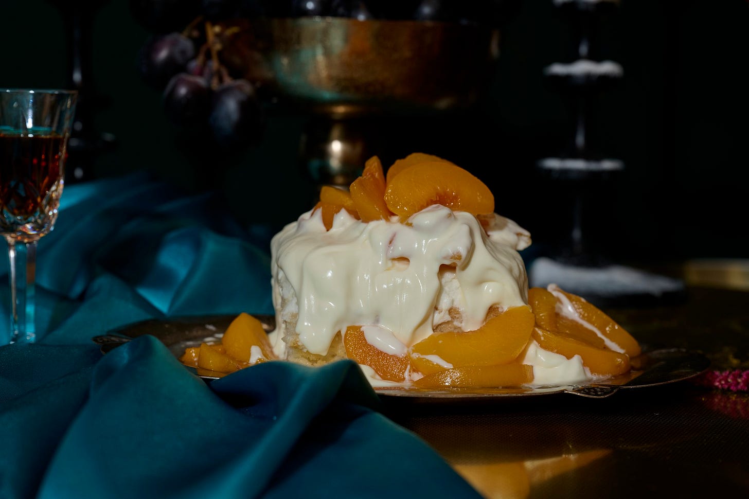 Close-up of a slice of Peaches & Cream Cake draped in whipped amaretto cream and topped with golden syrup-soaked peaches, styled on a vintage silver tray with a teal satin cloth and dark grapes in the background.