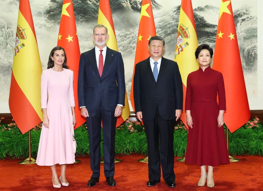 Four individuals stand in a formal group on a red carpeted area with green plants in front surrounded by flags of Spain and China against a backdrop of Chinese mountains and mist King Felipe VI in a blue suit and red tie stands beside Queen Letizia in a pink dress Xi Jinping in a dark suit and blue tie stands next to Peng Liyuan in a red dress all positioned formally with hands at sides