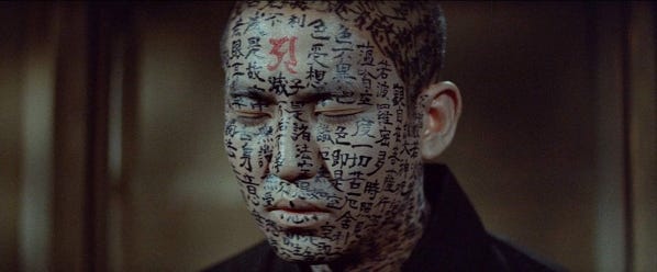 Film still from the 1964 horror anthology Kwaidan. A close-up of a monk’s face, eyes closed. His face is covered in black brush calligraphy in kanji, with one red character on his forehead that is larger than the others. His ears do not have any writing on them.