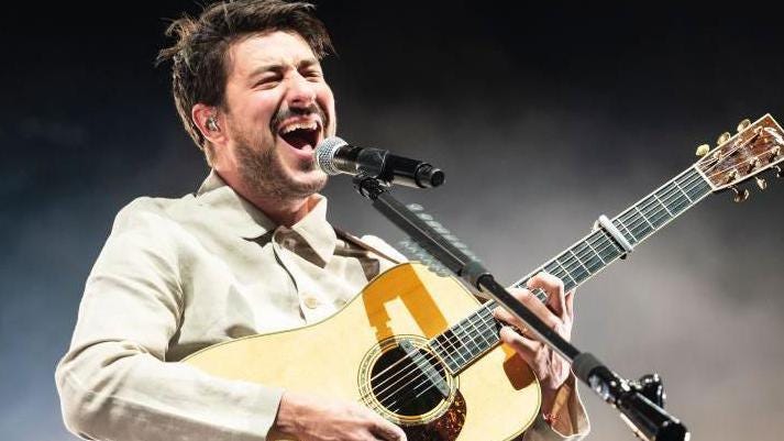 Marcus Mumford, wearing a light cream shirt, holding a guitar with his mouth open and a black and silver microphone in front of him