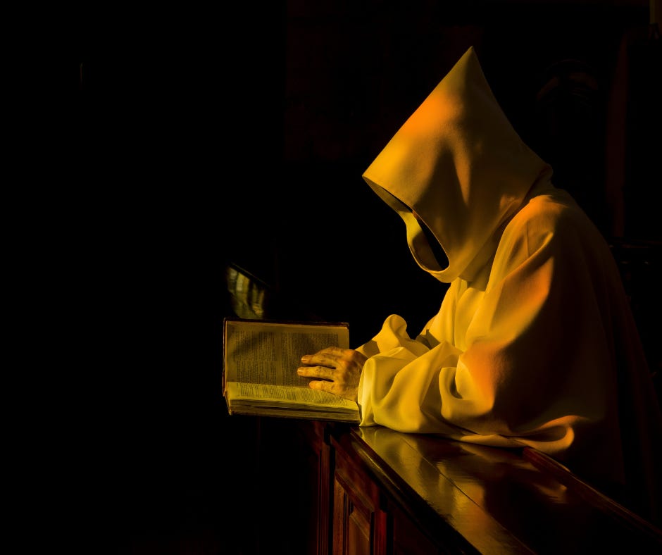 Alt text: A hooded figure in golden light reading an ancient book in near darkness.