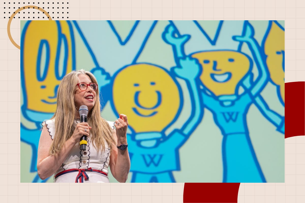 Bernadette Meehan, in red glasses and a white dress, speaks into a microphone in front of a giant backdrop of cheering blue-and-yellow cartoon Wikimedians. Bernadette Meehan, in red glasses and a white dress, speaks into a microphone in front of a giant backdrop of cheering blue-and-yellow cartoon Wikimedians.