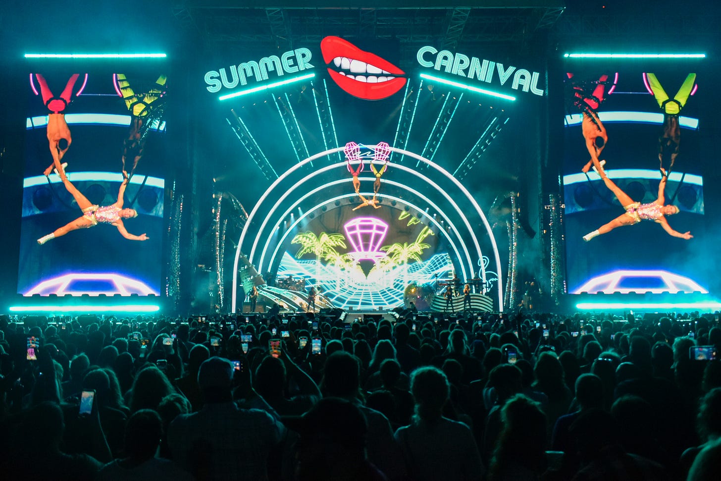 Two backup dancers hang by their feet from a prop several feet above the stage while holding P!nk by one arm and one leg.