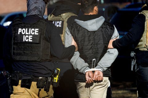 people in black longsleeved shirts and tactical vests bearing patches that read 'police ice' hold the arms of a man between them with his hands cuffed behind his back