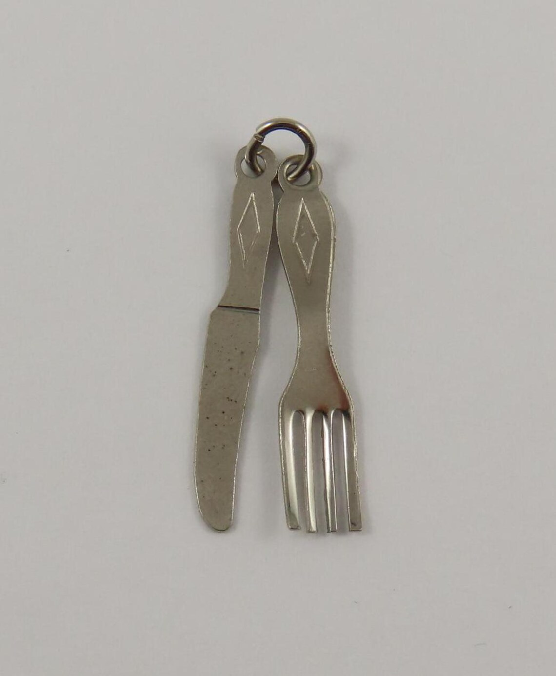 May include: A silver charm in the shape of a knife and fork. The charm has a diamond shape engraved on the handle of each utensil. May include: A silver charm in the shape of a knife and fork. The charm has a diamond shape engraved on the handle of each utensil.