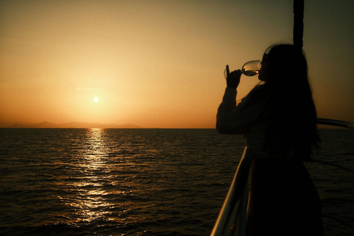 Man sieht die Umrisse einer Frau mit Weinglas in der Hand, die an einem Geländer steht und den Sonnenuntergang betrachtet. Man sieht die Umrisse einer Frau mit Weinglas in der Hand, die an einem Geländer steht und den Sonnenuntergang betrachtet.