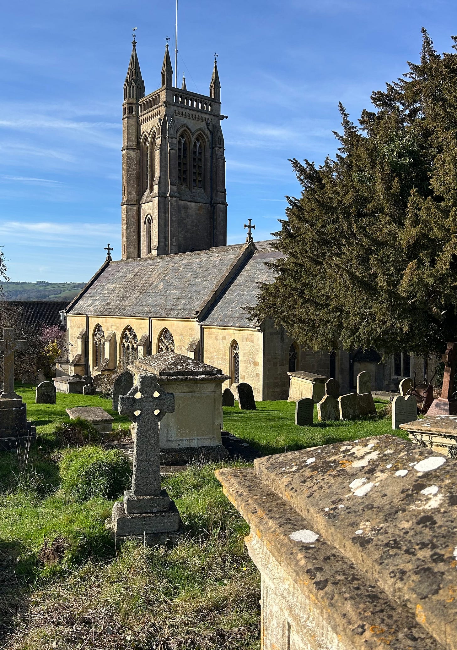 St Swithun’s Church, Bathford bathed in sunshine under a blue sky with wisps of high-level cloud. The tower overlooks the lanscape leading towards the city of Bath. In the foreground are graves and a tree. St Swithun’s Church, Bathford bathed in sunshine under a blue sky with wisps of high-level cloud. The tower overlooks the lanscape leading towards the city of Bath. In the foreground are graves and a tree.