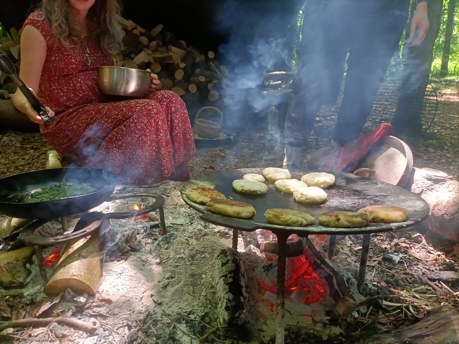 woman in red dress seated to right of campfire making flat breads on a metal tray suspended over the flames.