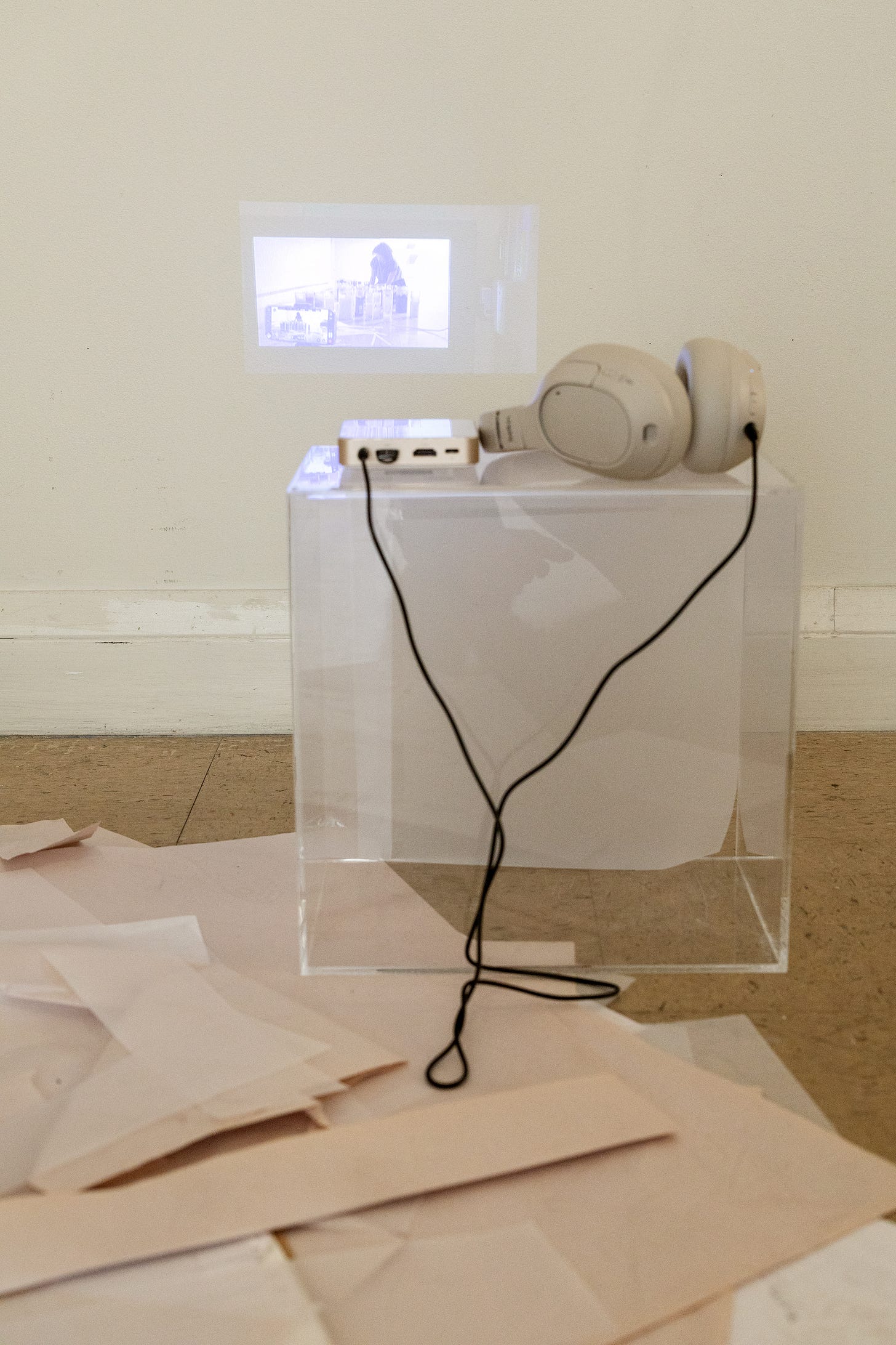 A clear acrylic vitrine on a gallery floor holds a small white projector and a pair of cream over-ear headphones, connected by a black cable. Loose sheets of newsprint paper are scattered on the floor around it.